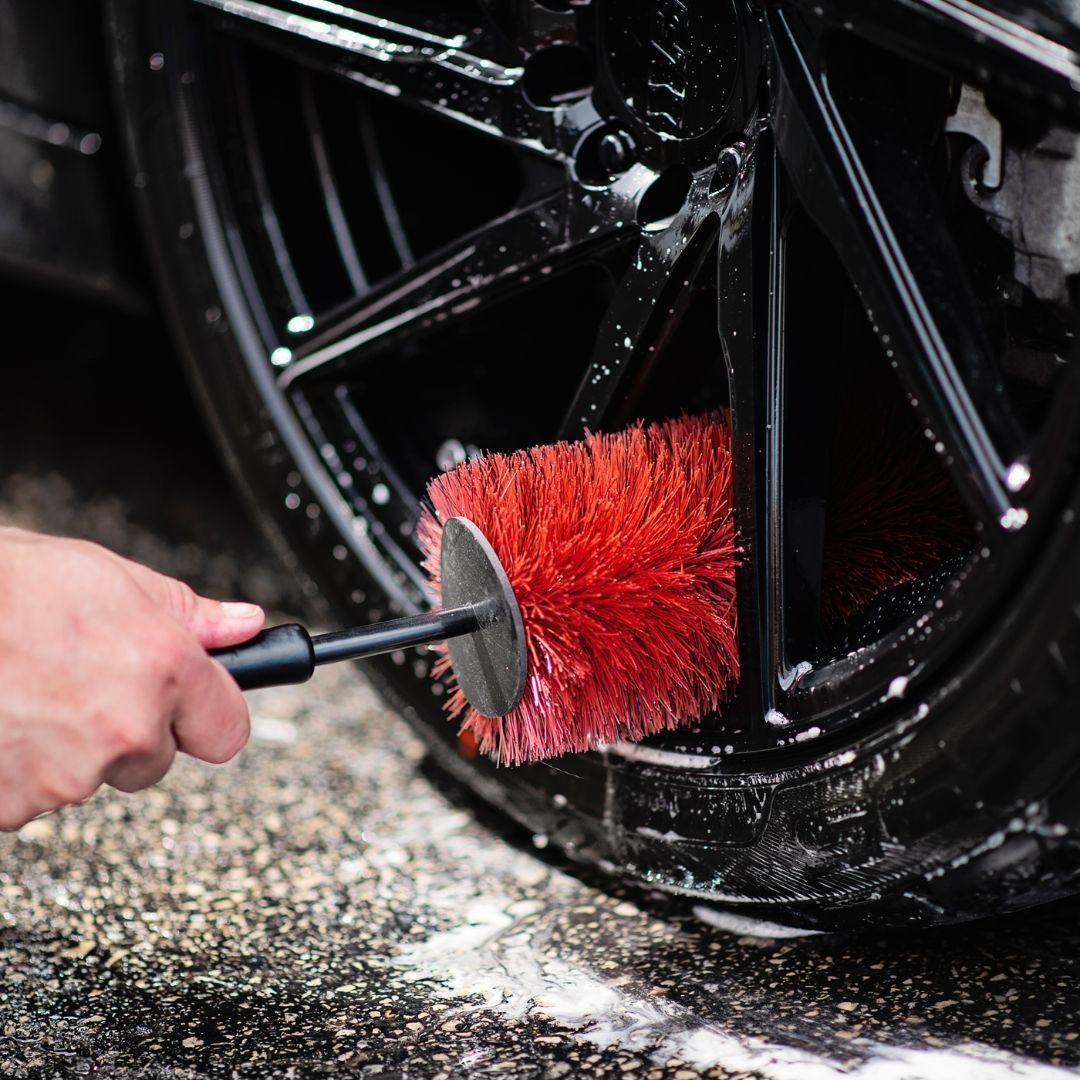 Person cleaning a black car wheel with a red circular brush attachment under water spray