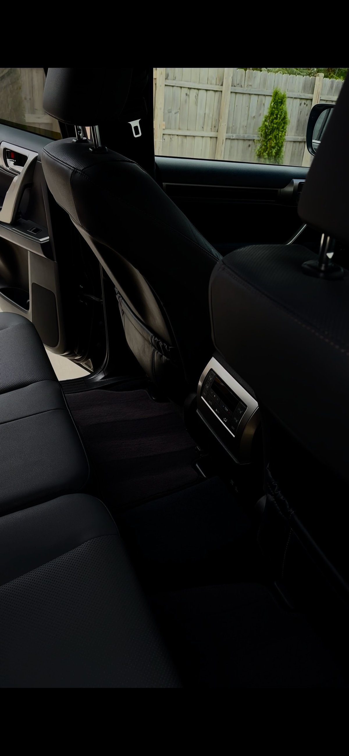 Interior view of a cars rear seat area with black upholstery, showing an open door and wooden fence visible through the window