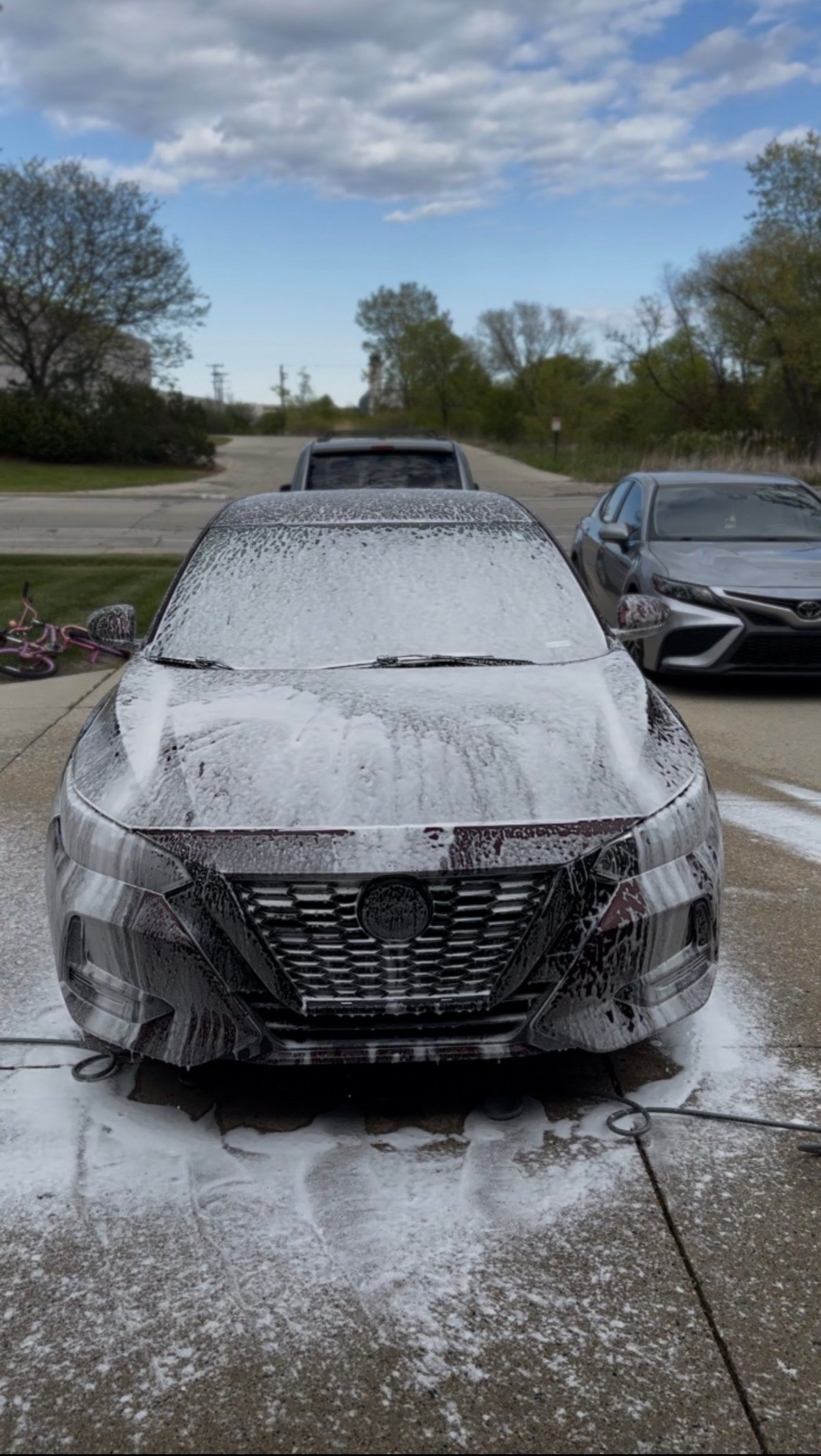 Silver sedan covered in soapy foam during outdoor car wash in parking lot with trees and other vehicles visible in background