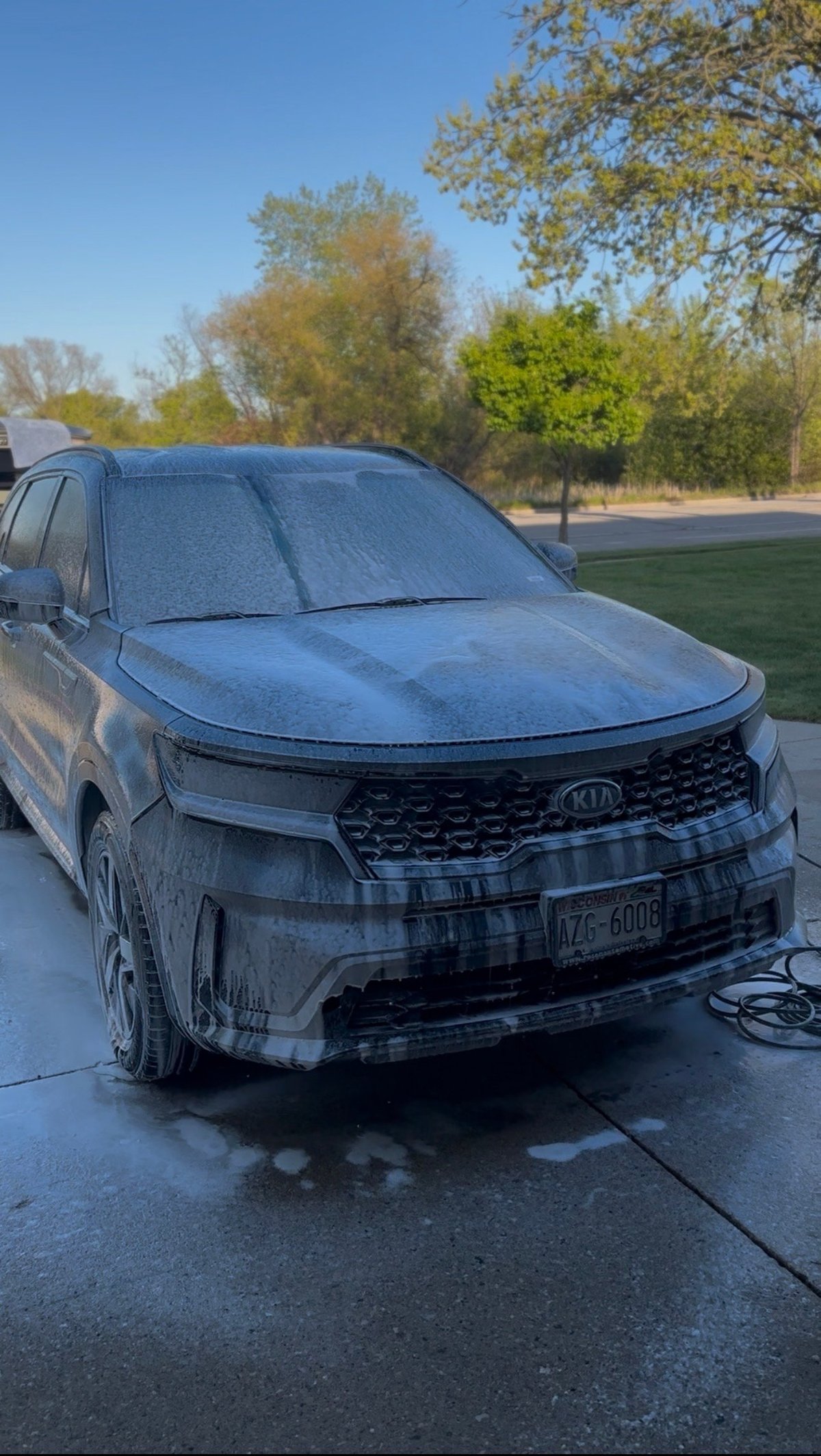 A blue Kia SUV covered in soap suds during an outdoor car wash on a driveway with trees in the background